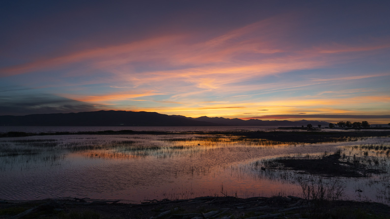 A wide view of the lake with yellow and pink streaky clouds above, the water reflects mostly pink and purple